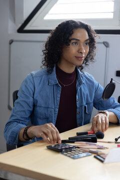 Non-binary adult sitting at wood desk under skylight arranging makeup tools and microphone in denim