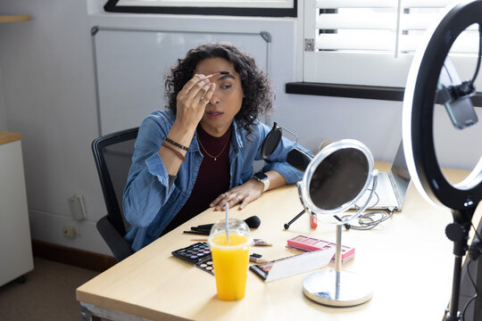 Adult non-binary person applying makeup with cotton pad at light wood desk with mirror, ring light