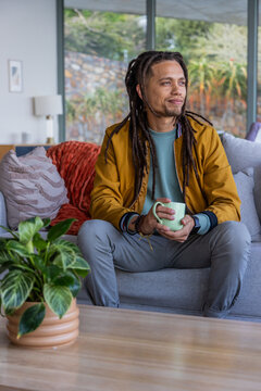 African American man wearing mustard coat sitting on sofa holding green mug looking out window