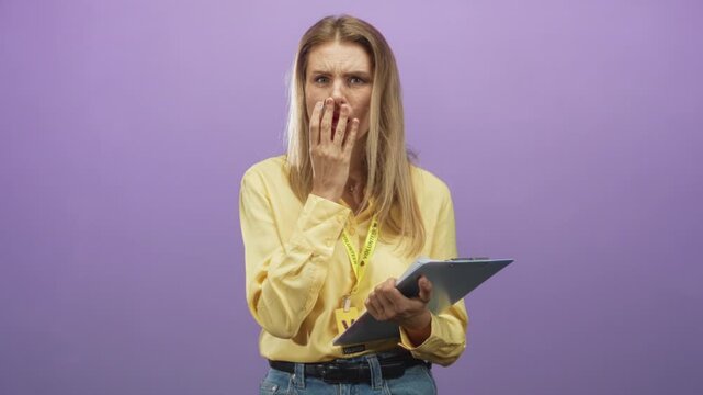 Woman covering mouth with hand while holding clipboard and visitor card, wearing yellow shirt and lanyard against a purple studio wall; concern doubt.