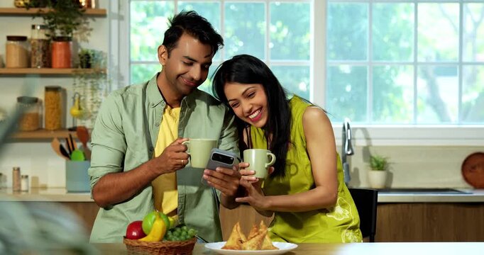 Indian asian young husband and wife having coffee at kitchen platform and surfing smartphone in joyful atmosphere in modern Indian home while sharing digital content and laughing during weekend