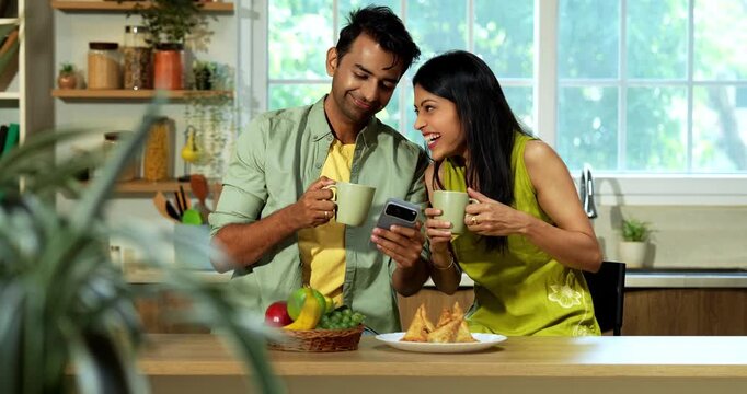 Indian asian young husband and wife having coffee at kitchen platform and surfing smartphone in joyful atmosphere in modern Indian home while sharing digital content and laughing during weekend