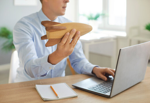 Cropped shot of company employee in shirt sitting at desk, using laptop and holding wooden rocket model in one hand. Focused businessman working on startup or business strategy for business growth.