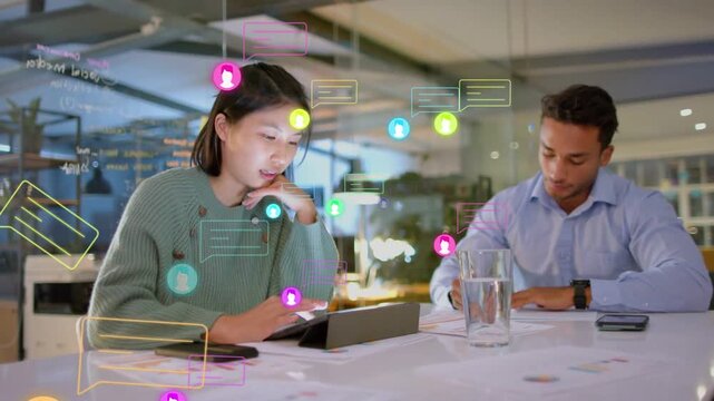 Woman tapping tablet and sending chat bubbles overlaying table while man writing notes for business
