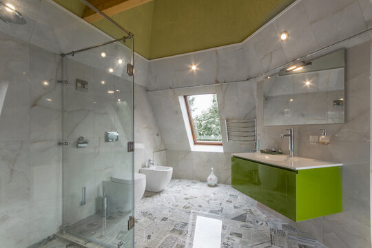 A bathroom shows a shower with clear glass walls, a sink with a green cabinet, and a window that lets in daylight. The floor features different patterns