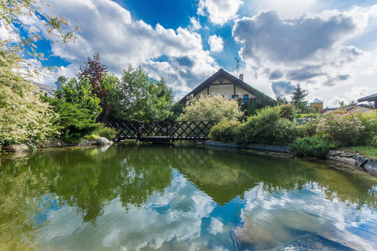 A house stands by a pond reflecting the sky and clouds. Trees surround the water, and a wooden bridge crosses over the pond. It is afternoon with clear skies