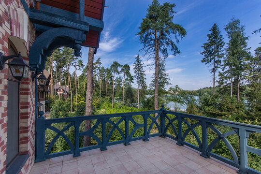A balcony overlooks a forest filled with tall trees. A lake can be seen in the distance under a clear blue sky. The scene represents a natural setting during the day