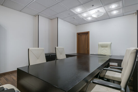 A meeting room shows a large dark table surrounded by empty white chairs. The walls are plain and there is a wooden door. The lights above provide even illumination in the space