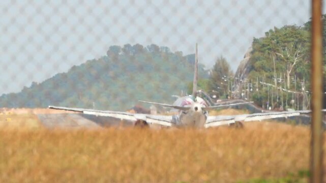Old airplane rests behind chain-link fence. Tall, dry grass surrounds the aircraft. Green hill rises behind the field. No people visible in this scene