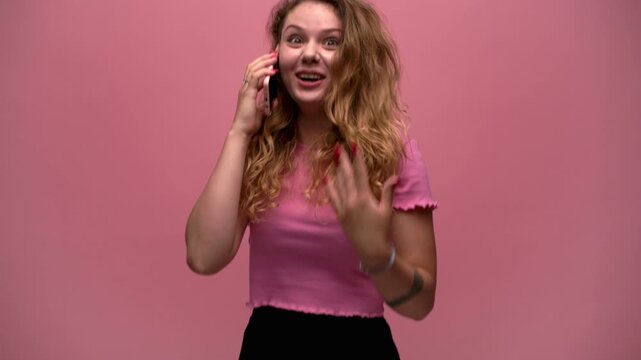 Overjoyed young woman with curly hair talking on smartphone receiving good news and celebrating on pink background