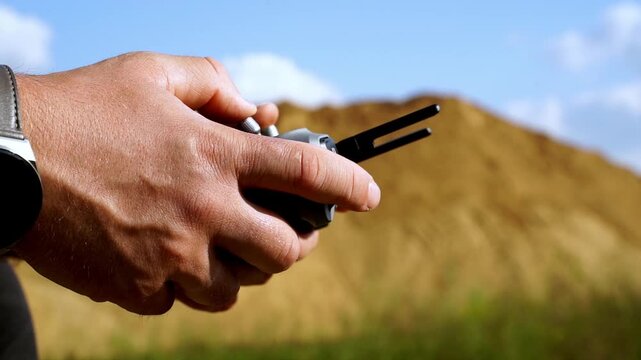 Hands grip a remote control firmly. Sky and earth form a blurred backdrop. Dirt mound stands still behind the hands. Focus rests on the controller's buttons. User prepares for drone flight control