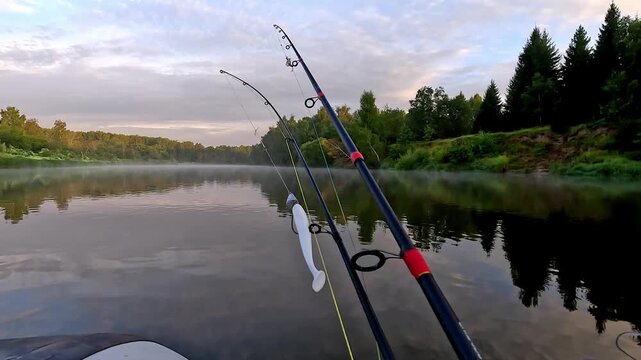 Calm river reflects trees and sky. Fishing rods rest in a quiet boat. Mist drifts across the surface. Early morning stillness prevails. Natural peace invites viewers to pause