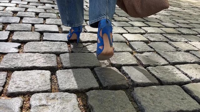 walking legs blue high heels cobblestone street woman in denim jeans and stiletto sandals walking away from camera on stone pavement outdoors concept of fashion, urban lifestyle, footwear