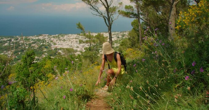 Woman walks up a flower lined trail overlooking Capri town and coastline below in summer sunny day