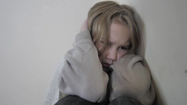 Young girl in strong stress sits on floor in corner afraid of something. Child covers ears to not hear parents quarrel. Family conflicts as problem for child psyche. Fear and panic of child.
