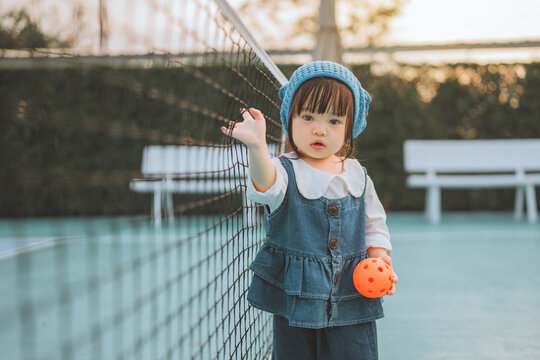 Cute Asian toddler girl holding an orange ball on a pickleball court at sunset. Happy child in denim outfit learning sports, promoting active lifestyle, wellness, and childhood development