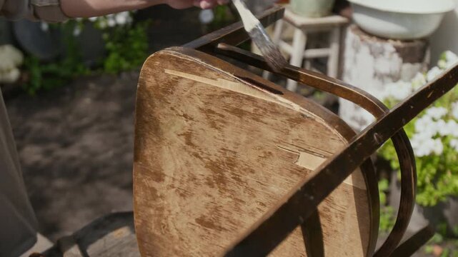 Medium shot of hands of anonymous rural man painting old chair with wood stain using brush, while restoring vintage furniture in garden in summer, leading sustainable simple life in countryside