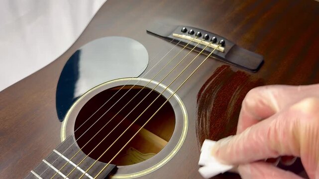 Close-up of a person wearing gloves polishing a wooden acoustic guitar body with a white cloth.