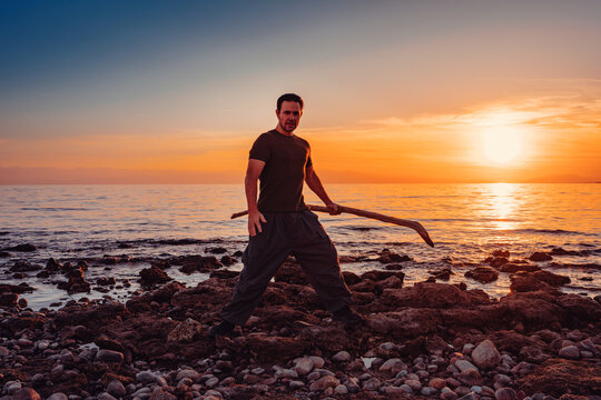 Man with wooden stick posing on rocky beach at sunset