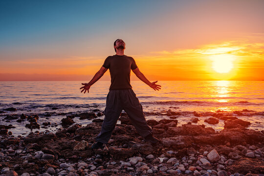 Man standing on rocky seashore at sunset with arms wide open