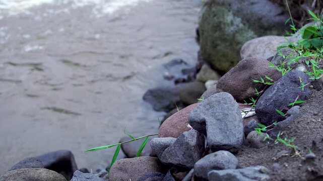 Close up of natural volcanic rocks on the riverbank of Mount Kelud lahar flow path in Indonesia, with a murky river flowing in the blurred background.
