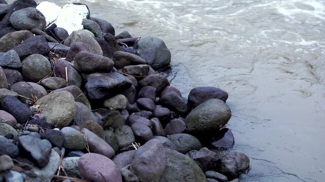 Dark rounded volcanic rocks piled on a riverbank beside flowing murky water in the Mount Kelud lahar path, Indonesia.