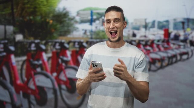 Man holding smartphone and giving thumbs up by a row of red rental bicycles on street; joy travel convenience.