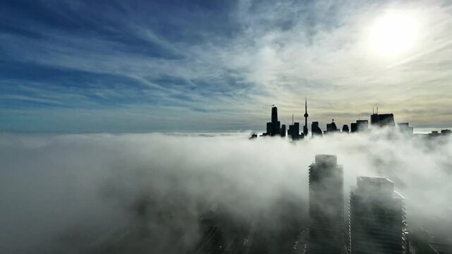 Cinematic aerial drone footage of downtown Toronto covered in dense morning fog, with sunlight piercing through the mist and creating a glowing, almost ethereal atmosphere. Modern high-rise buildings