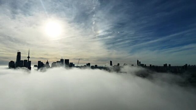 Cinematic aerial drone footage of downtown Toronto covered in dense morning fog, with sunlight piercing through the mist and creating a glowing, almost ethereal atmosphere. Modern high-rise buildings