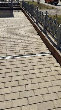 Metal grate spillway on paving stones white downpipe above, patterned metal cover set into paver joints, thin rust streaks.