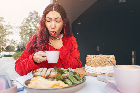 Young woman experiencing pain and choking while drinking hot coffee at an outdoor restaurant during breakfast time