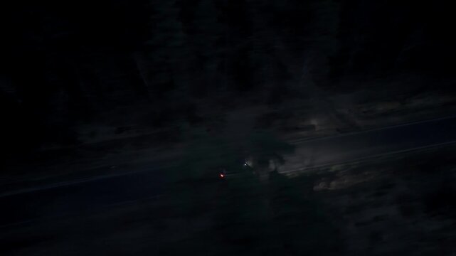 Night aerial footage of a single car crossing dense forest with lights on recorded from high altitude tracking shot