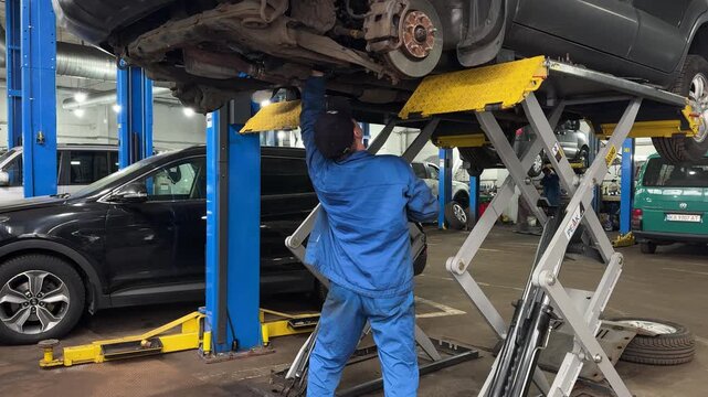 Car mechanic working under lifted vehicle in garage workshop during auto repair service. Brake maintenance, suspension inspection and professional vehicle service in automotive garage.