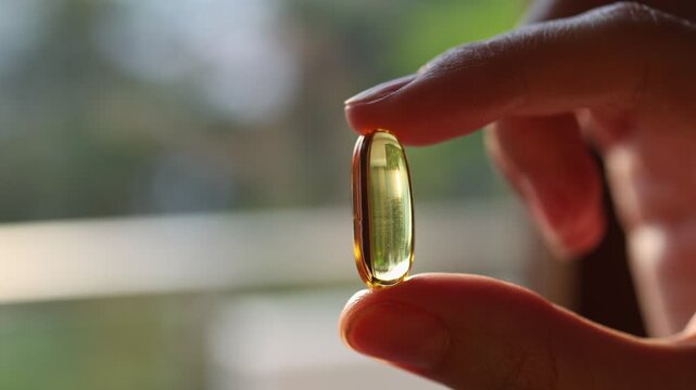 Macro shot of a hand holding a yellow fish oil capsule or vitamin D supplement against a blurred background. Close-up of a golden transparent softgel pill. Health and omega-3 stock footage.