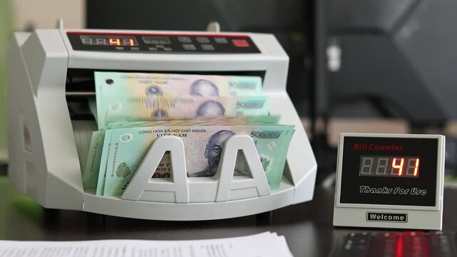 Closeup shot of banknote counting machine with Vietnamese Dong money (VND). Automatic currency counter equipment processing paper bill with Ho Chi Minh portrait vignette. Finance industry in Vietnam.