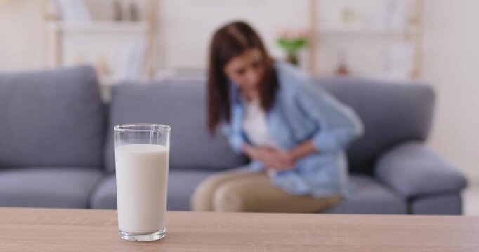 Glass of cow milk in focus as woman with lactose intolerance and food allergy holding belly due to stomach pain and bad digestion after drinking. Girl with food allergy sitting on sofa at home
