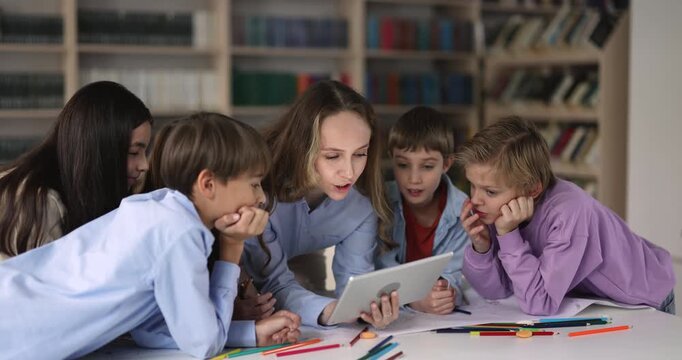 Female teacher and group of primary school children watching online content gathered around digital tablet at classroom table. Learning, technology, application use during art class activity