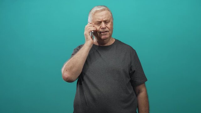 Man holding smartphone to ear, talking with furrowed brow and grey hair, wearing dark tshirt against teal studio backdrop, mid chest frontal pose; concern isolation.