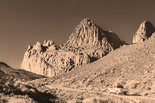 Monochrome sepia-toned Hoggar desert mountain range in Tamanrasset, Algeria. A white pickup truck drives along the road in the foreground, across the rugged, sunlit terrain under a clear sky.