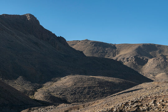 Looking up toward the Assekrem mountain, the small stone building of Father de Foucauld's hermitage is visible at the distant summit. The rugged, sun-drenched slopes contrast with deep shadows in the 