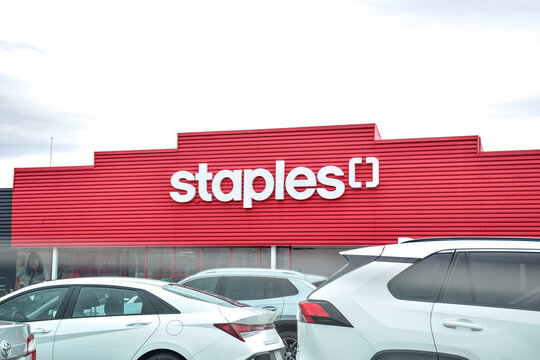 Staples store entrance viewed from plaza parking lot. Retail storefront with signage visible in commercial shopping area highlighting office supply shopping. Aurora, Ontario, Canada, April 2026