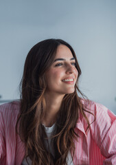 Fototapeta premium Smiling young woman with long brown hair looks to the side, radiating relaxed confidence and warmth in a soft, casual indoor lifestyle portrait. Natural expression, modern fashion and positive mood.
