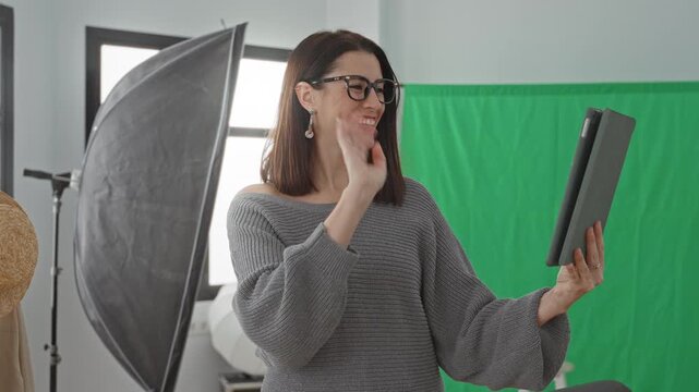 Woman holding a tablet and waving in a studio with green screen and softbox lighting while smiling at the screen; happiness connection creativity.