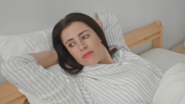 Woman lying on bed in striped pajama top, hand behind head, smiling, pillow and wooden frame visible in building; relaxed contentment.