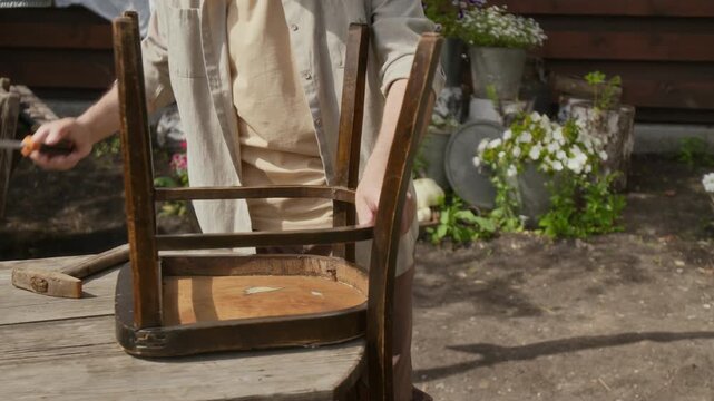 Medium midsection shot of unrecognizable male village dweller repairing old furniture outdoors in summer, checking chair and fixing legs with hammer, while busy with routine domestic work