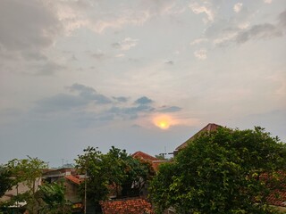 Obraz premium Sunset Over Residential Area with Warm Evening Light, Warm sunset view over houses and trees with soft clouds in the sky, creating a peaceful evening atmosphere. 