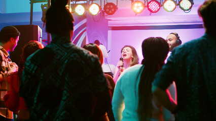 Fototapeta premium Happy woman doing karaoke at party, singing songs with DJ in front of friends on dance floor. Confident girl having fun with live show perfromance next to electronic mixing station at nightclub.