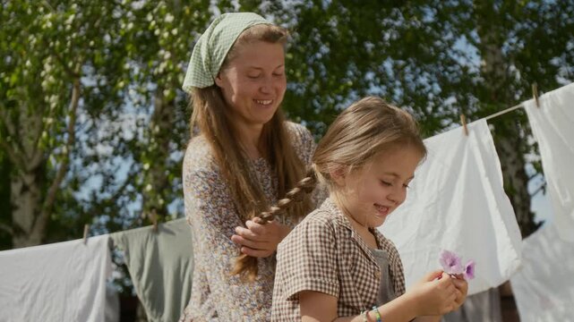 Medium shot of loving rural mom in peasant dress and headscarf styling long hair in braid for young daughter and chatting, while little girl is playing with flowers, laundry drying on lines