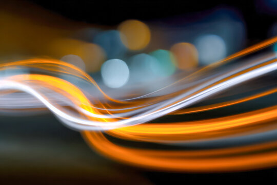 Dynamic flowing orange and white light trails creating a vibrant wave pattern against a dark blurred background with colorful bokeh effects at night