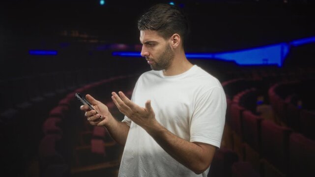 Man holding smartphone with raised hand questioning gesture in a movie theater building, white tshirt and beard visible; confusion.